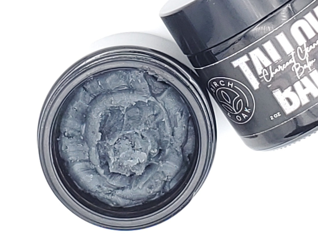 A jar of Tallow Charcoal Cleansing Balm with its lid removed, showing the dark gray balm inside. The product is labeled with the brand name and the words 'Charcoal Cleansing Balm'.